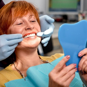 An older woman with red hair admires her smile in the mirror after receiving her dental implants in Huntington Beach